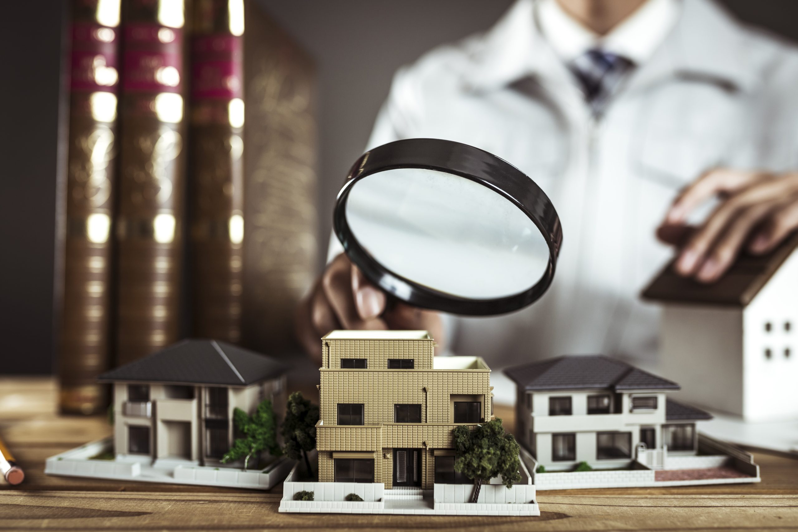 白衣の専門家（鑑定士）が虫眼鏡で住宅模型を細部まで調査・鑑定しているイメージ画像（擁壁調査の専門性を表現）